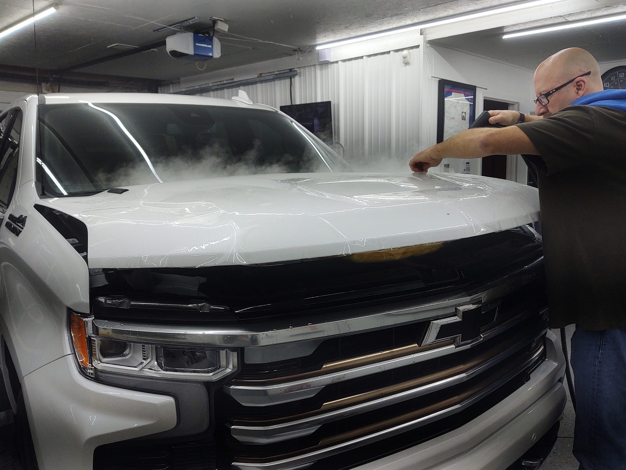PPF being steamed onto white Silverado hood, dramatic steam and clear film visible